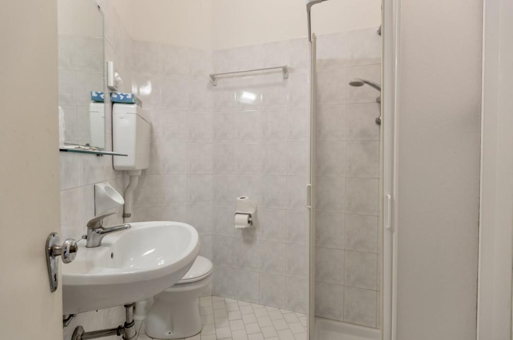Bagno semplice, piastrelle bianche, doccia piccola, lavabo e WC, atmosfera pulita e ordinata.