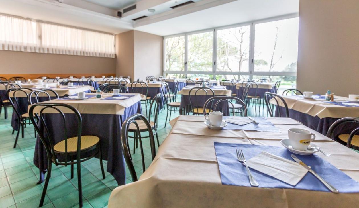 Sala da pranzo luminosa, tavoli apparecchiati con tazze, posate e tovagliette blu.