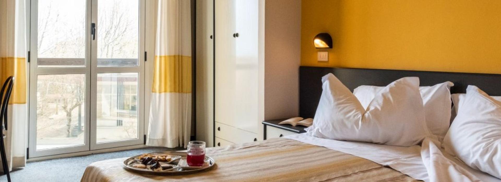 Camera accogliente con letto soffice, valigia rossa, colazione sul vassoio e vista luminosa.